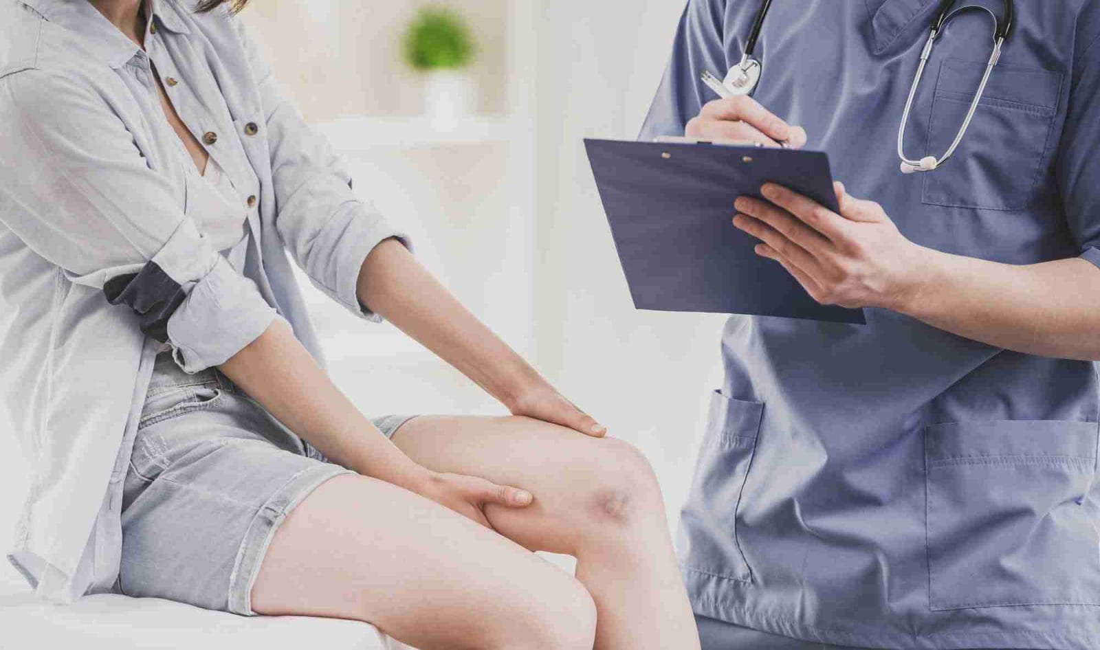 Doctor taking notes on a clipboard while a patient describes her knee pain during a consultation.