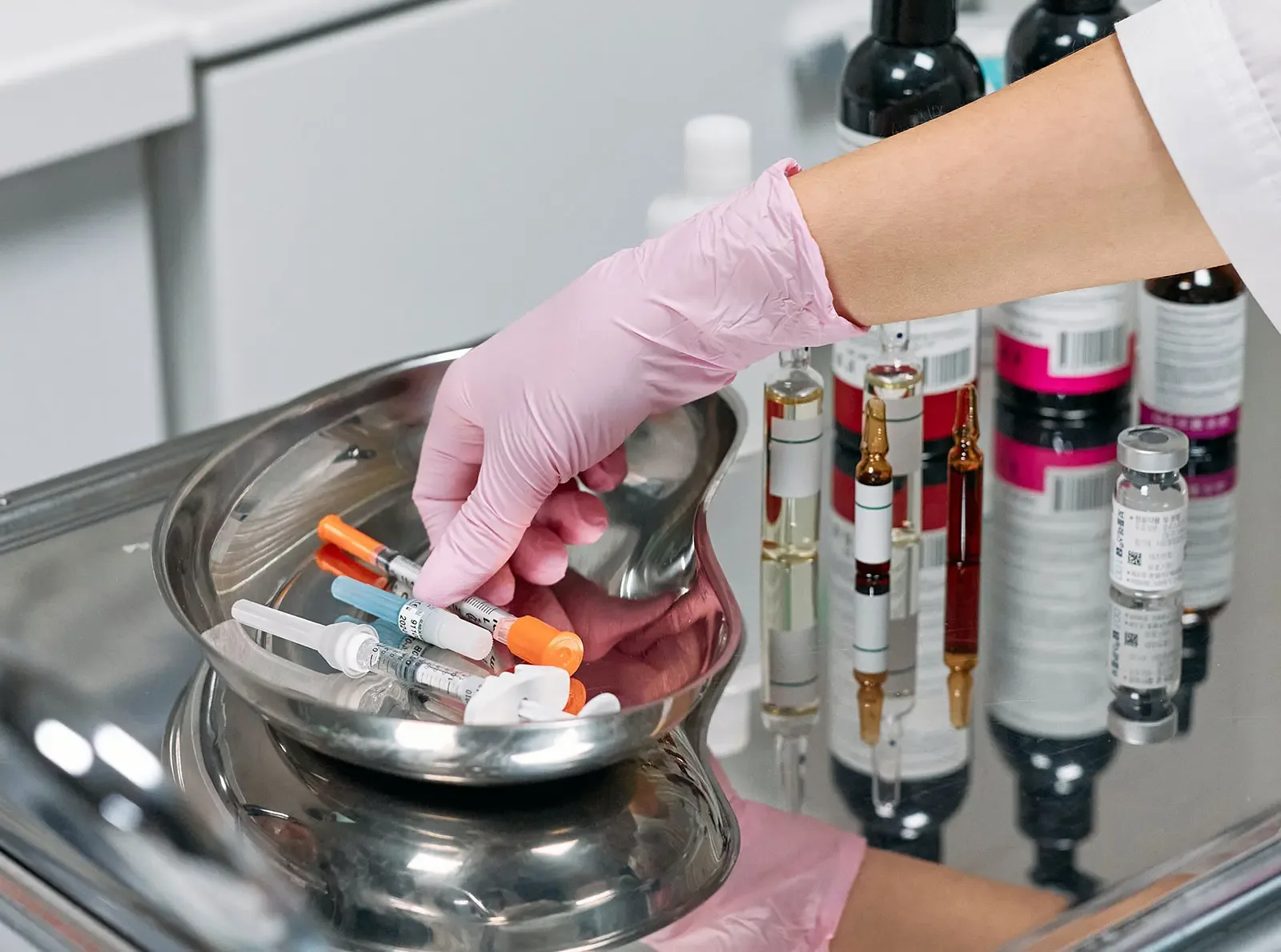 Gloved clinician selecting syringes from a stainless tray of injection supplies and medication vials.