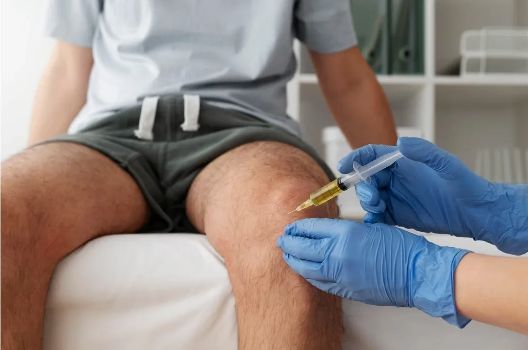 A gloved clinician gives a patient an injection into the knee joint in a clinic exam room.