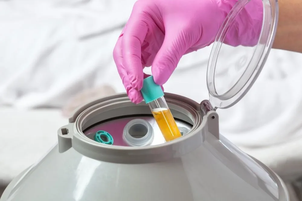A gloved hand places a tube of golden platelet-rich plasma into a centrifuge.