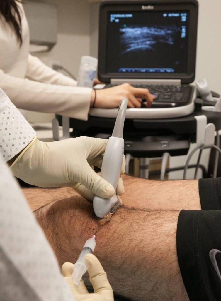 A gloved clinician positions an ultrasound probe on a patient's knee to guide a joint injection.