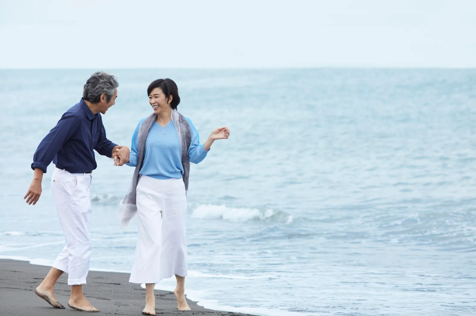 A smiling older couple holds hands while walking barefoot along the ocean shore.