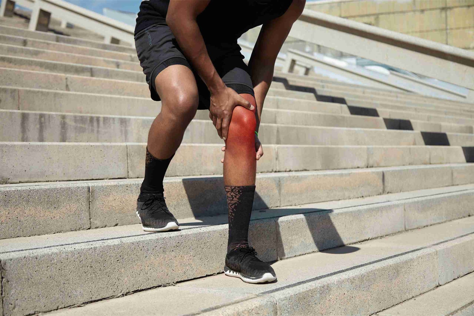 Athlete pausing on outdoor stairs and clutching his knee, which is highlighted red to indicate pain.