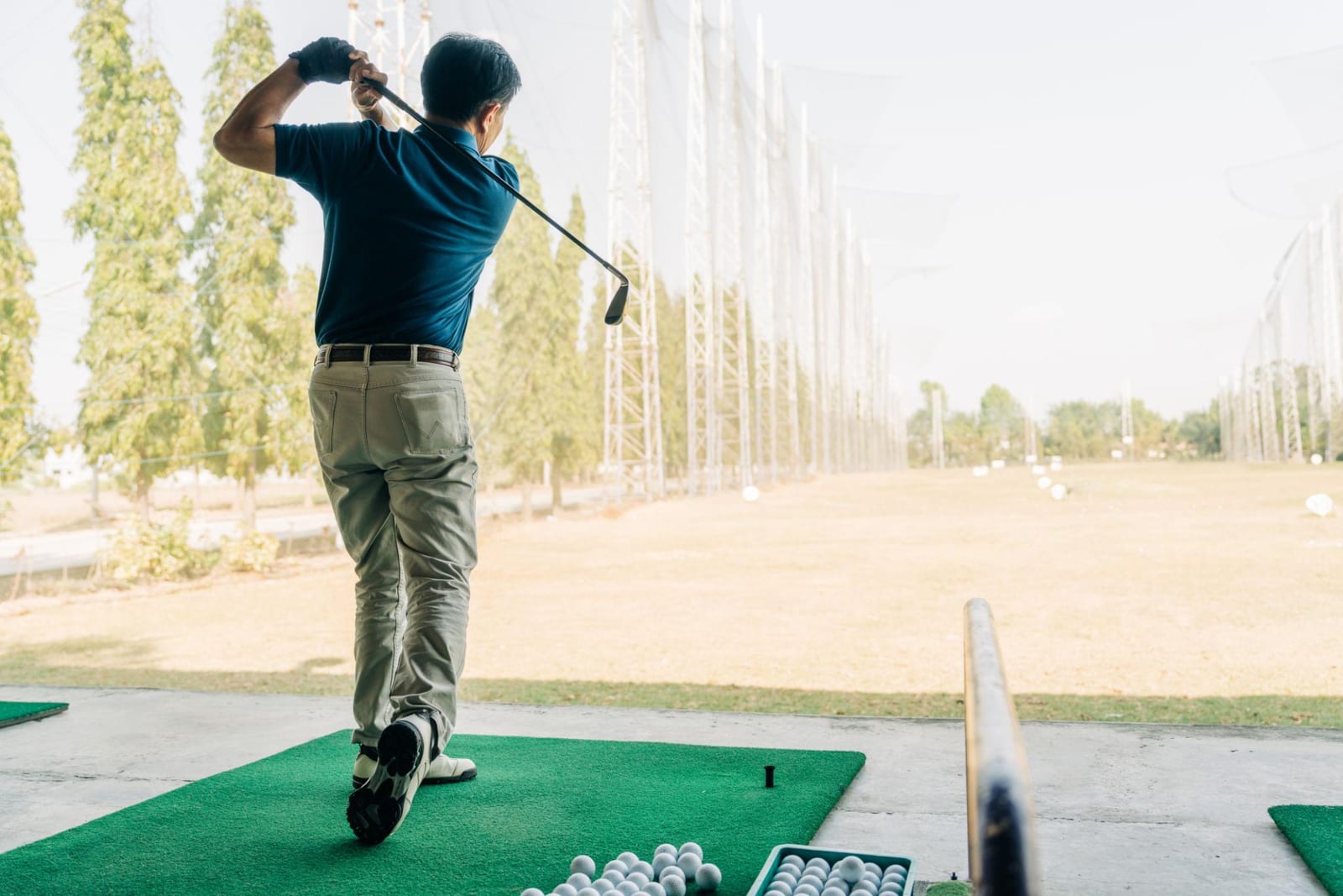 Older man in a blue polo finishing a golf swing at a driving range.