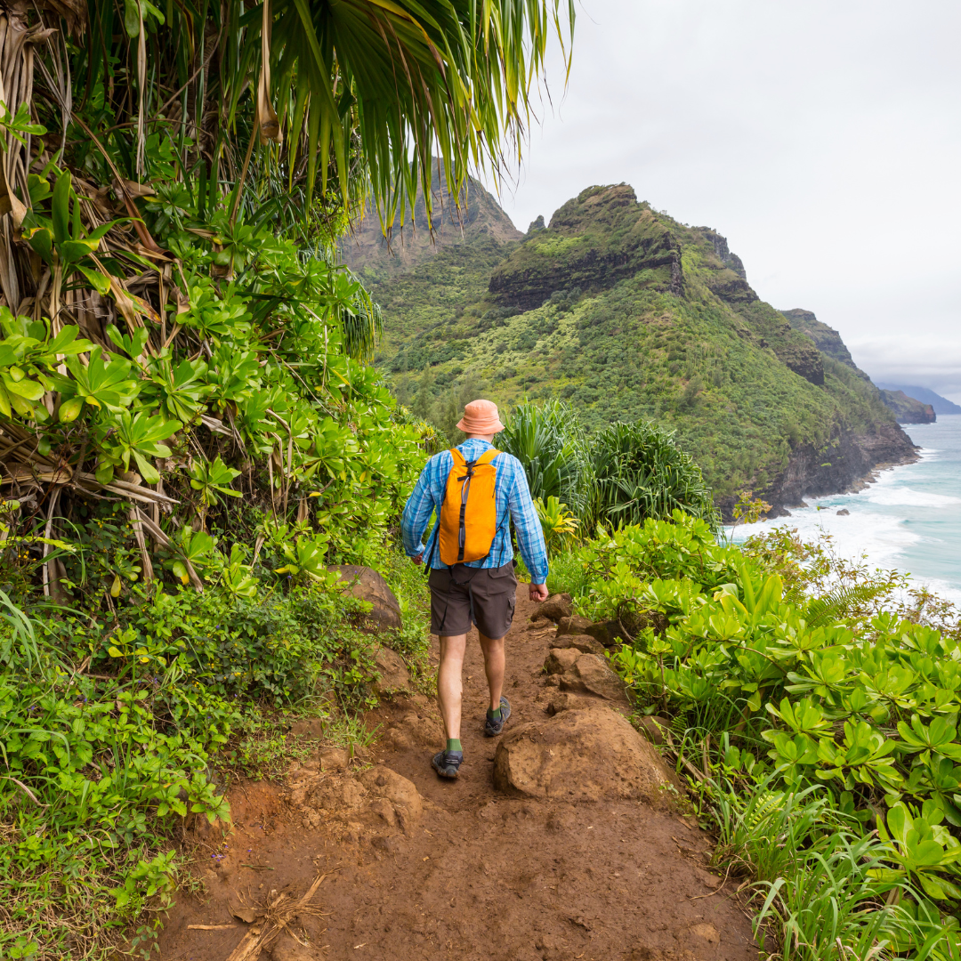 A hiker with an orange backpack walks the Kalalau Trail along Kauai's Nā Pali Coast.