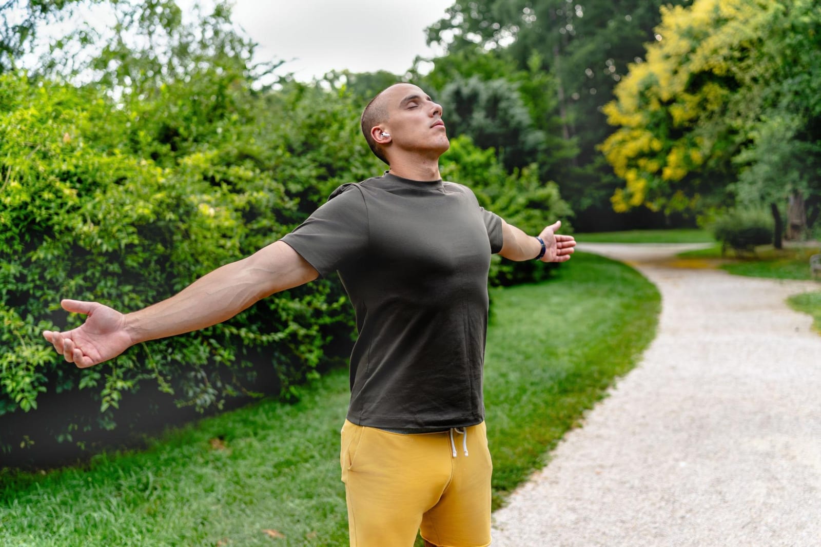 Man standing on a park path with arms outstretched, breathing deeply outdoors.