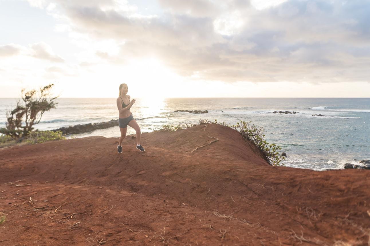 A runner jogs along a red-dirt coastal bluff above the ocean in Hawai'i at sunrise.