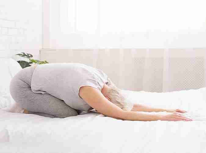 Older woman stretching in child's pose on a bed, a gentle mobility exercise for the back and hips.