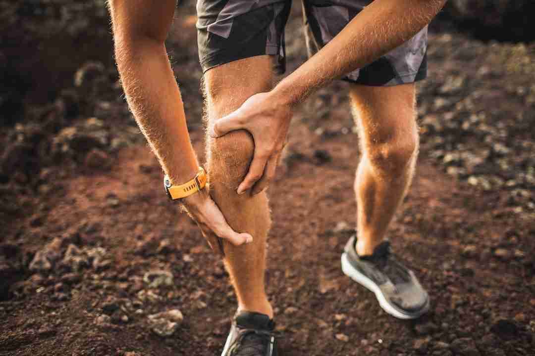 A trail runner grips his knee in pain on a red-dirt Hawai‘i path — activity-related pain in a previously injured knee is an early sign of post-traumatic arthritis.