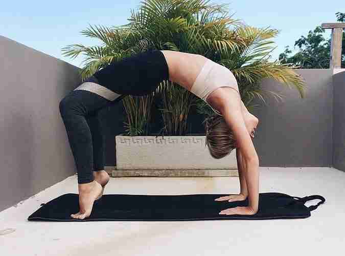 Woman holding bridge pose on a yoga mat in a palm-lined outdoor courtyard, a hip-strengthening posture.
