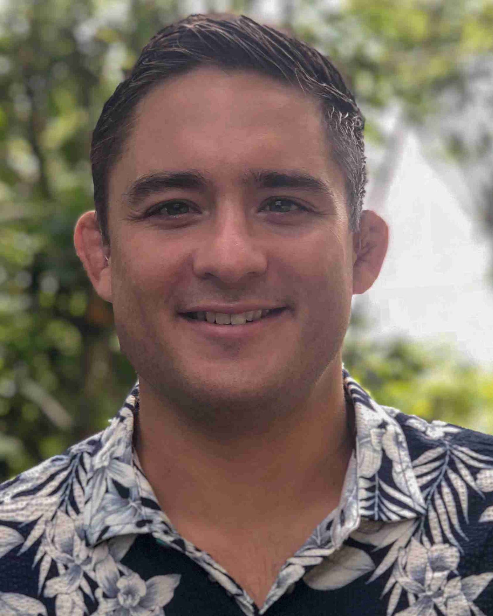 Dr. Paul Norio Morton, MD, smiling outdoors in an aloha shirt with tropical greenery behind him in Hawai‘i.