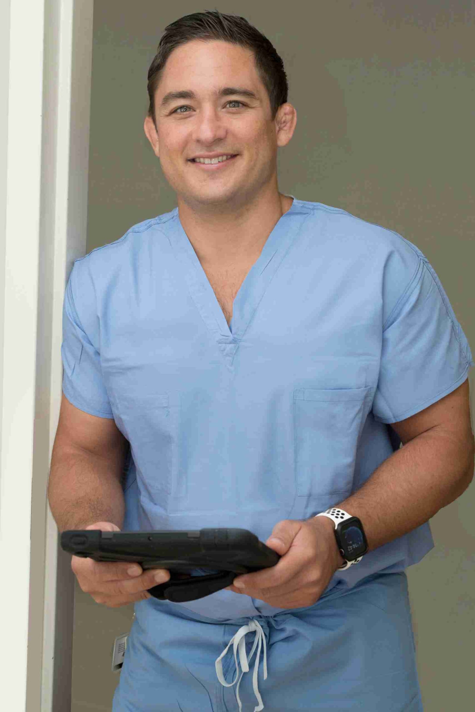 Dr. Paul Morton in blue scrubs holding a tablet — he sees patients from every Hawaiian island and across the Pacific by video visit.