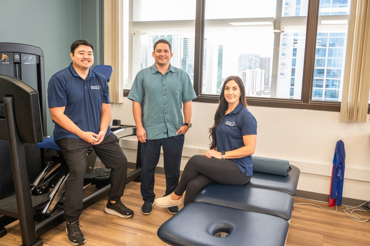 PB&J Physical Therapy team of three in the Honolulu treatment room with exercise equipment and city views.