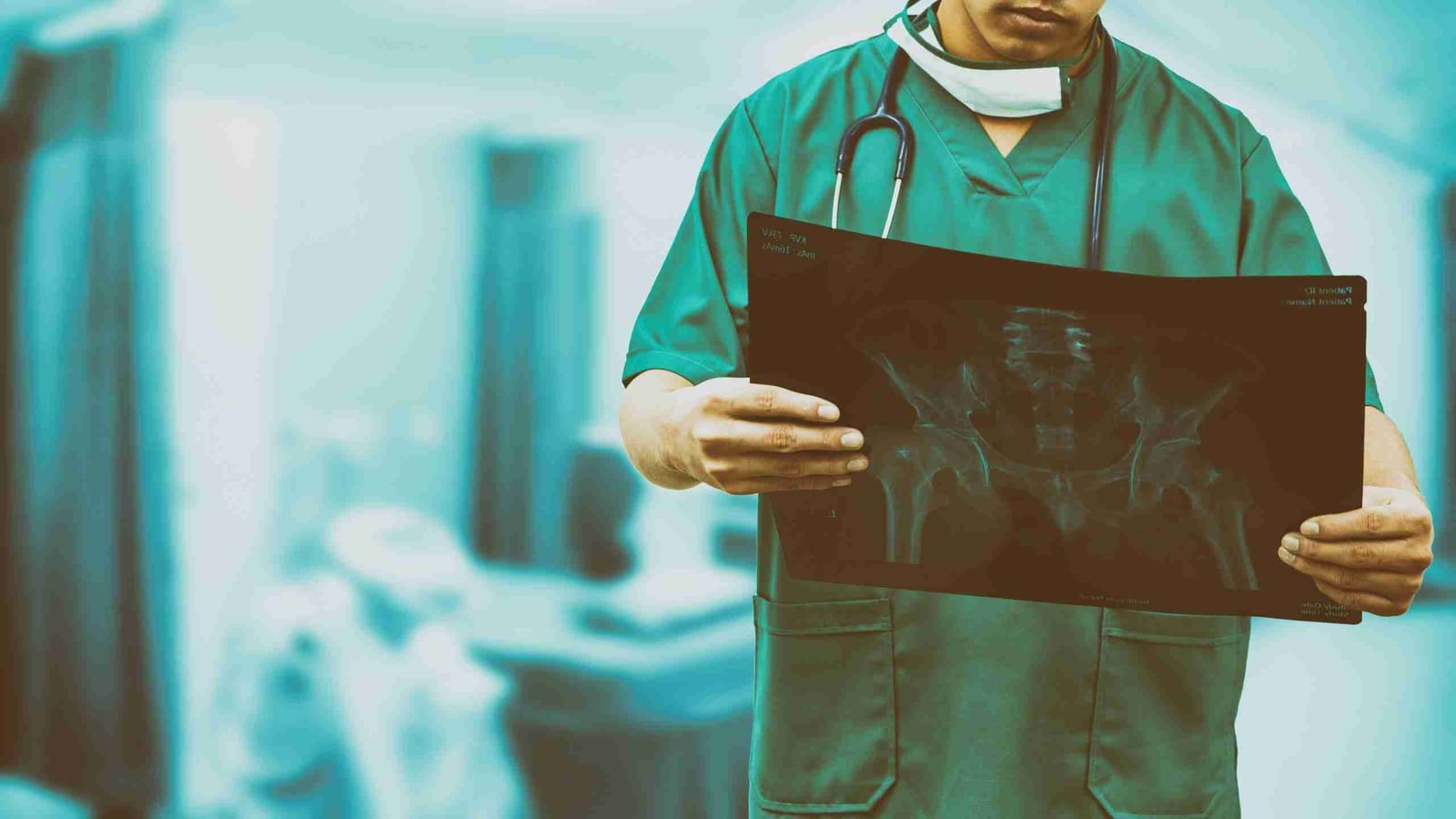 An orthopedic surgeon in green scrubs reviews a pelvis X-ray film in a hospital corridor.